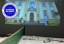 Olympische Spiele x Actif/Réactif / Kulturforum Paris Das Foto zeigt einen Tischtennistisch, der mit Papier zum darauf malen ausgelegt ist sowie zwei Tischtennisschläger. Im Hintergrund läuft eine Videoübertragung zu einem Tischtennismatch. Der Malgrund auf dem Tischtennistisch ist bereits bemalt.