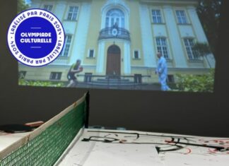 Olympische Spiele x Actif/Réactif / Kulturforum Paris Das Foto zeigt einen Tischtennistisch, der mit Papier zum darauf malen ausgelegt ist sowie zwei Tischtennisschläger. Im Hintergrund läuft eine Videoübertragung zu einem Tischtennismatch. Der Malgrund auf dem Tischtennistisch ist bereits bemalt.