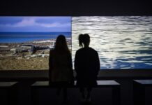 On the Road Again 2 – “Living Investment Risk” / Kulturforum Peking Das Bild zeigt zwei Personen von Hinten die auf einer Bank sitzend zwei Bilder bei einer Ausstellung von Oliver Ressler betrachten