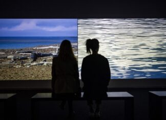 On the Road Again 2 – “Living Investment Risk” / Kulturforum Peking Das Bild zeigt zwei Personen von Hinten die auf einer Bank sitzend zwei Bilder bei einer Ausstellung von Oliver Ressler betrachten