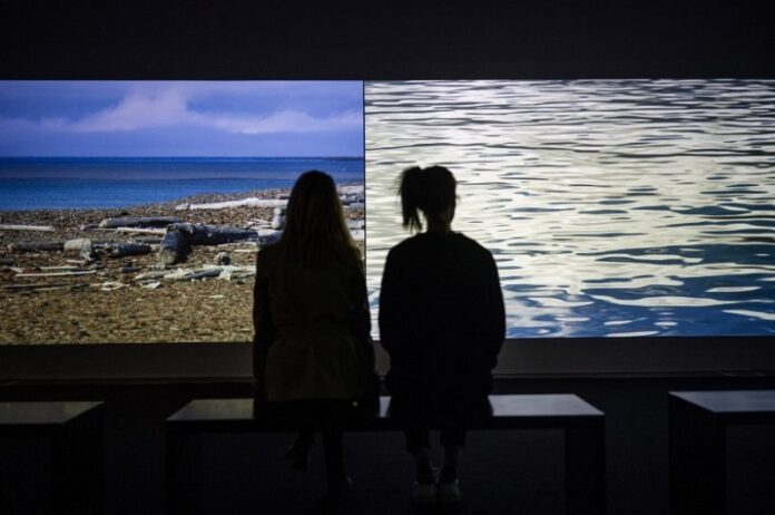 Das Bild zeigt zwei Personen von Hinten die auf einer Bank sitzend zwei Bilder bei einer Ausstellung von Oliver Ressler betrachten