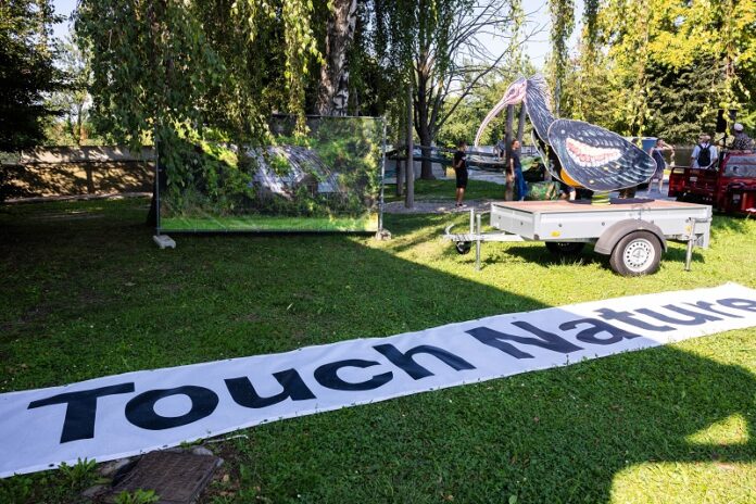 Das Bild zeigt ein Transparent mit der Aufschrift Touch Nature, eine Fotografie mit einem überwucherten Gewächshaus und die Skulptur eines Waldrapps auf einem Wagen.