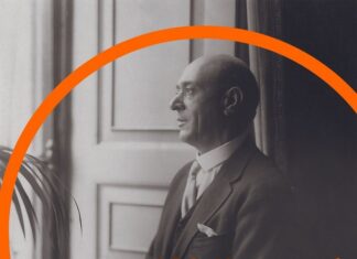 150 Years of Arnold Schönberg! / Kulturforum London Das Bild zeigt Arnold Schönberg, der am Fenster steht und hinausblickt. Im Bild ist ein oranger Bogen als Designelement sichtbar, unter dem der Schriftzug „150 Years of Arnold Schönberg“ in Orange und Weiß zu sehen ist.