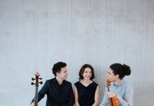 Ausstellungseröffnung: Arnold Schönberg – 150 Years & Concert by Trio Callas / Kulturforum New York Das Bild zeigt drei Musiker sitzend vor einer grauen Wand, die sich gegenseitig ansehen und dabei lächeln. Zwei der Musiker haben ihre Instrumente bei sich, ein Cello und eine Geige.