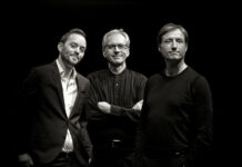 John Arman Organ Trio in Polen / Kulturforum Warschau Das Bild zeigt drei Männer, die sich vor einem dunklen Hintergrund befinden. - Der Mann links hat kurzes Haar, trägt ein helles Hemd und darüber ein dunkles Sakko. Er neigt seinen Kopf leicht zur Seite und lächelt freundlich in die Kamera. - Der Mann in der Mitte hat graues Haar und trägt eine Brille sowie ein dunkles, langärmliges Hemd. Er steht mit verschränkten Armen und einem entspannten Gesichtsausdruck da. - Der Mann rechts hat ebenfalls kurzes Haar und trägt ein einfaches, schwarzes Langarmshirt. Sein Blick ist direkt auf die Kamera gerichtet, und er zeigt ein dezentes Lächeln. Das Bild ist in Schwarz-Weiß gehalten, was den Fokus auf die Gesichter und Ausdrücke der Männer lenkt. Die Beleuchtung hebt die Personen klar vom dunklen Hintergrund ab und sorgt für starke Kontraste.