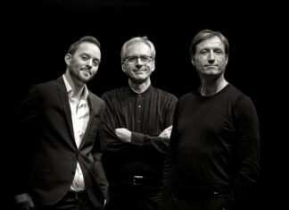 John Arman Organ Trio in Polen / Kulturforum Warschau Das Bild zeigt drei Männer, die sich vor einem dunklen Hintergrund befinden. - Der Mann links hat kurzes Haar, trägt ein helles Hemd und darüber ein dunkles Sakko. Er neigt seinen Kopf leicht zur Seite und lächelt freundlich in die Kamera. - Der Mann in der Mitte hat graues Haar und trägt eine Brille sowie ein dunkles, langärmliges Hemd. Er steht mit verschränkten Armen und einem entspannten Gesichtsausdruck da. - Der Mann rechts hat ebenfalls kurzes Haar und trägt ein einfaches, schwarzes Langarmshirt. Sein Blick ist direkt auf die Kamera gerichtet, und er zeigt ein dezentes Lächeln. Das Bild ist in Schwarz-Weiß gehalten, was den Fokus auf die Gesichter und Ausdrücke der Männer lenkt. Die Beleuchtung hebt die Personen klar vom dunklen Hintergrund ab und sorgt für starke Kontraste.