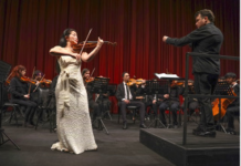 CHILE Tournee Tomoko Mayeda / Botschaft Santiago de Chile Das Bild zeigt die Geigerin Tomoko Mayeda mit einem silbernen Kleid vor dem Orchester, rechts von Ihr der Dirigent auf einem Stehpult.