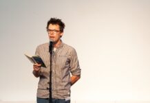 Poetry Slam + Workshop / Botschaft Usbekistan Das Bild zeigt Markus Köhle stehend hinter einem Mikrophon mit einem Buch in der Hand bei einer Lesung.