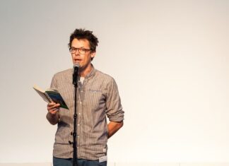 Poetry Slam + Workshop / Botschaft Usbekistan Das Bild zeigt Markus Köhle stehend hinter einem Mikrophon mit einem Buch in der Hand bei einer Lesung.