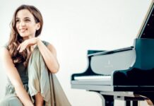 Klavierkonzert von Mitra Kotte / Kulturforum Budapest Das Bild zeigt die sitzende Pianistin Mitra Kotte lächelnd nach rechts schauend links neben einem schwarzen Konzertflügel vor einem hellen Hintergrund.