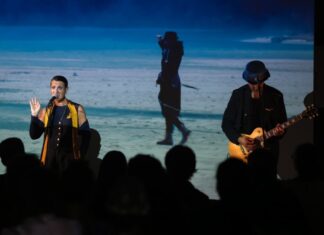 Orlando Trip / Kulturforum Rom Das Bild zeigt einen Musiker, der auf einer Bühne E-Gitarre spielt sowie eine Sängerin hinter dem Mikrofon. Im Hintergrund ist ein projiziertes Bild einer schwarz gekleideten Person am Strand zu sehen. Im Vordergrund verfolgt das Publikum die Aufführung.