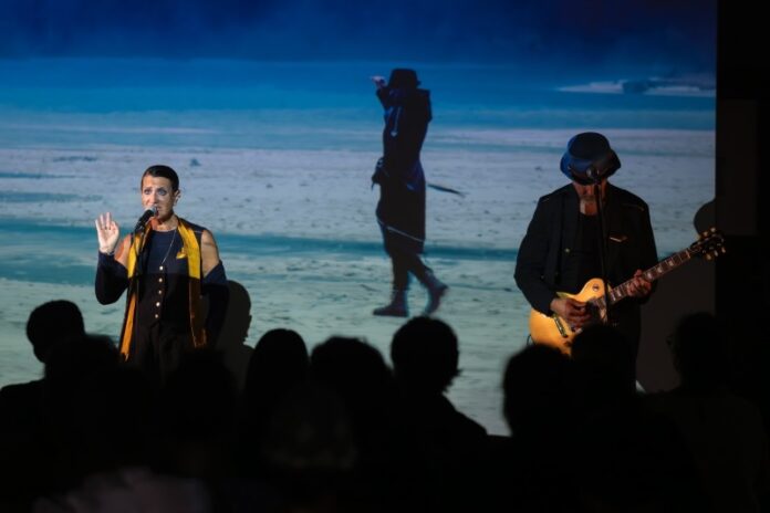 Das Bild zeigt einen Musiker, der auf einer Bühne E-Gitarre spielt sowie eine Sängerin hinter dem Mikrofon. Im Hintergrund ist ein projiziertes Bild einer schwarz gekleideten Person am Strand zu sehen. Im Vordergrund verfolgt das Publikum die Aufführung.