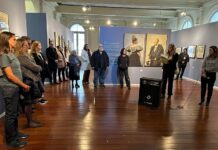 Architekturausstellung / Botschaft Brasilia Das Bild zeigt einen taubenblau gestrichenen Ausstellungsraum mit Objekten aus der Otto Wagner Ausstellung, davor eine Menge an Besuchende im Halbkreis stehend und rechts Ges.-BR Judith Schildberger bei Ihren Begrüßungsworten.