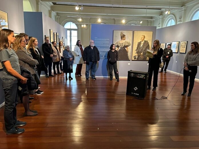 Das Bild zeigt einen taubenblau gestrichenen Ausstellungsraum mit Objekten aus der Otto Wagner Ausstellung, davor eine Menge an Besuchende im Halbkreis stehend und rechts Ges.-BR Judith Schildberger bei Ihren Begrüßungsworten.