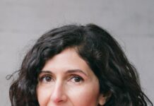 Literatur² mit Nava Ebrahimi / Botschaft Oslo Das Bild zeigt die Autorin Nava Ebrahimi vor grauem Hintergrund. Sie lächelt auf dem Foto, hat dunkles, gewelltes Haar, das ihr etwa bis zum Kinn reicht, braune Augen und trägt einen pinken Strickpullover mit Rollkragen.