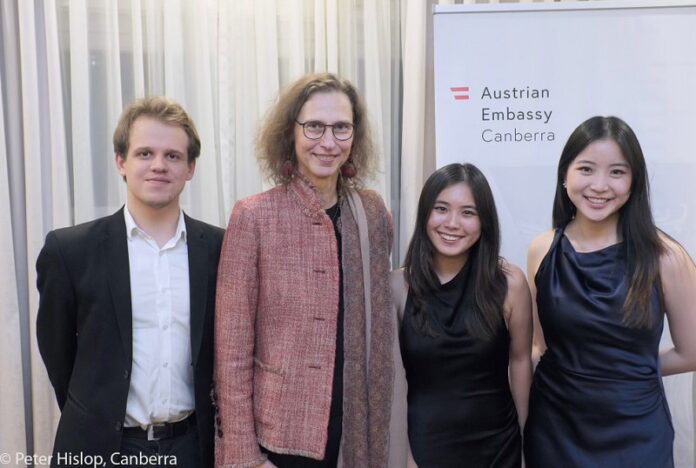 Das Bild zeigt Botschafterin Kögler mit dem Trio Alpenrose vor einem Logo der Österreichischen Botschaft Canberra