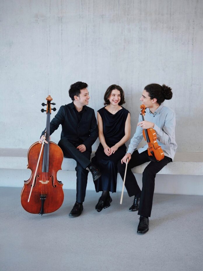 Das Bild zeigt das Trio Callas: den Cellisten Lucas Garcia Muramoto mit seinem Instrument, die Klavierspielerin Bella Schütz und den Violinisten Miguel Rocha mit seiner Geige.