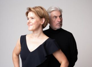 Viktor Ullmann und Rainer Maria Rilke / Kulturforum Berlin Das Bild zeigt die Pianistin Senka Brankovic und den Sprecher Helmut Mooshammer in einer Halbtotalen, beide von der Hüfte aufwärts, vor einem neutralen, hellen Hintergrund. Links im Bild steht die Pianistin mit kurzen, leicht gewellten, hellbraunen Haaren. Sie trägt ein schwarzes Oberteil und lächelt leicht, während sie nach links aus dem Bild schaut. Ihre Hände ruhen auf ihren Hüften, was ihr eine selbstbewusste und entspannte Haltung verleiht. Rechts von ihr steht ein Mann mit grauem Haar und Bart, der nach rechts blickt. Er trägt einen schwarzen Rollkragenpullover und hat einen nachdenklichen Gesichtsausdruck. Die beiden Personen sehen sich nicht an.