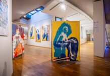 Ausstellung: OPUS 24 / Kulturforum Berlin Das Bild zeigt eine moderne Kunstausstellung in der Galerie des Österreichischen Kulturforums Berlin. Im Vordergrund befindet sich ein großes Gemälde mit einer stilisierten Darstellung einer Frau. Die Farben des Gemäldes sind hauptsächlich Gelb und Blau. Links davon ist ein weiteres Kunstwerk zu sehen, das eine stehende Frau in einem roten Kleid darstellt. Weiter hinten an der Wand hängen drei Porträts von Frauen in ähnlichem Stil, mit kontrastreichen Farben wie Blau und Gelb. Die Siebdruck-Technik ist deutlich erkennbar und verleiht den Bildern eine rohe, handwerkliche Ästhetik.