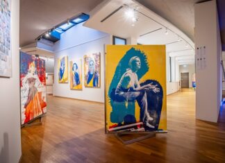 Ausstellung: OPUS 24 / Kulturforum Berlin Das Bild zeigt eine moderne Kunstausstellung in der Galerie des Österreichischen Kulturforums Berlin. Im Vordergrund befindet sich ein großes Gemälde mit einer stilisierten Darstellung einer Frau. Die Farben des Gemäldes sind hauptsächlich Gelb und Blau. Links davon ist ein weiteres Kunstwerk zu sehen, das eine stehende Frau in einem roten Kleid darstellt. Weiter hinten an der Wand hängen drei Porträts von Frauen in ähnlichem Stil, mit kontrastreichen Farben wie Blau und Gelb. Die Siebdruck-Technik ist deutlich erkennbar und verleiht den Bildern eine rohe, handwerkliche Ästhetik.