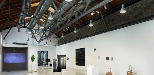 Das Bild zeigt die in weiß gehaltene Ausstellungshalle mit zahlreichen Ausstellungsstücken. Bilder, Fotografien, sowie Skulpturen