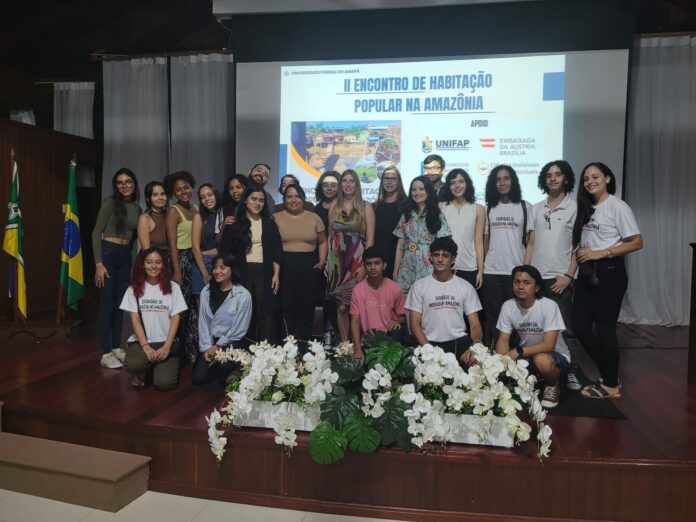 Das Bild zeigt eine Gruppe von 23 Personen, vor allem brasilianische Studierende, auf einer Bühne mit der Ankündigung der Veranstaltung im Hintergrund auf einer Leinwand eingeblendet.