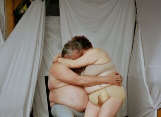 34. Internationales Festival „Monat der Fotografie 2024“ / Kulturforum Bratislava Das erste Foto zeigt zwei Menschen, eine Frau und einen Mann, die sich auf einem Stuhl sitzend innig umarmen. Die Frau sitzt auf dem Schoß des Mannes. Im Hintergrund befinden sich Leinentücher, die die Möbel des Raumes verdecken.