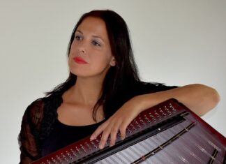 Konzert im Rahmen der Kampagne “Orange the World” / Kulturforum Bratislava Das Bild zeigt ein Foto der Hackbrettistin Hemma Pleschberger-Schmölzer. Sie hält das Hackbrett vor sich in Händen und blickt nach rechts aus dem Bild hinaus mit einem sehnsuchtsvollen Blick, ihre Lippen leuchten in hellem Rot und sie hat lange braune Haare.