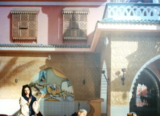 A Sea of Stories / Kulturforum Istanbul Das Foto zeigt das Werk „A Sea of Stories“ von Lisl Ponger. Die orientalische Szene zeigt vor einem Haus einen schlafenden Mann im Stuhl, vor sich eine Wasserpfeife. Hinter ihm sitzt eine dunkelhaarige Frau am Divan, rechts von ihm ein Mann mit einer Wasserpfeife in der Hand