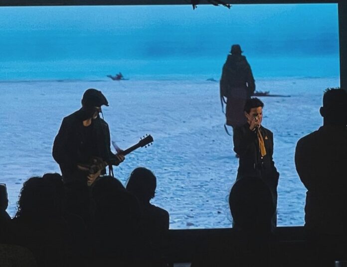 Das Bild zeigt die Aufführung in Colombo. Ein Gitarrenspieler und ein Sänger auf der Bühne vor einer Leinwand, die einen das Meer überquerenden Orlanda zeigt.