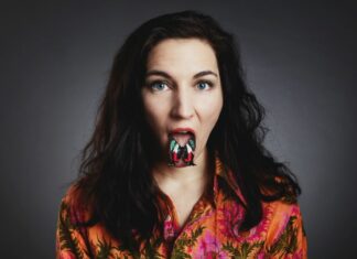 Konzert Anna Anderluh / Kulturforum Prag Das Bild zeigt eine weiße Frau ab den Schultern vor grauem Hintergrund. Sie trägt eine wild gemusterte Bluse in orange-, rosa-, und Grüntönen. Ihre braunen Haare reichen bis zu ihren Schultern, ihre blauen Augen und ihr rot geschminkter Mund sind weit geöffnet. Auf ihrer herausgestreckten Zunge liegt ein schwarzer Schmetterling mit roten, weißen und türkisen Akzenten auf seinen ausgebreiteten Flügeln.