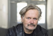 Ed. Hauswirth / Kulturforum Rom Das Bild zeigt ein Porträt des Künstlers Ed.Hauswirth: er trägt eine schwarze Lederjacke und Jeans. Sitzend auf einer Stufe hat er die Hände vor sich locker verschränkt. Die Haare sind blond und etwass länger, er schaut direkt mit offenen Blick in die Kamera.