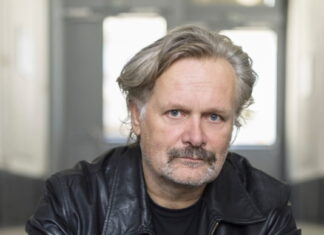 Ed. Hauswirth / Kulturforum Rom Das Bild zeigt ein Porträt des Künstlers Ed.Hauswirth: er trägt eine schwarze Lederjacke und Jeans. Sitzend auf einer Stufe hat er die Hände vor sich locker verschränkt. Die Haare sind blond und etwass länger, er schaut direkt mit offenen Blick in die Kamera.