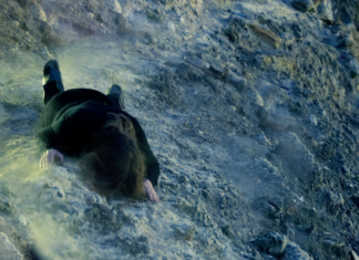 Shaken Grounds / Kulturforum Rom Das Bild zeigt eine Person, die im dunklen Anzug und in Bauchlage auf den Phlegräischen Feldern liegt.