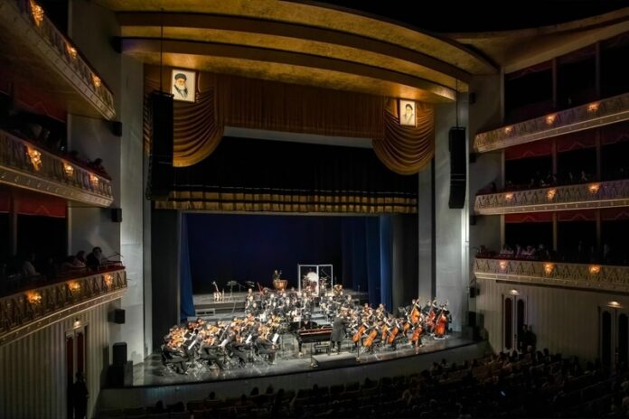 Das Bild zeigt die Teheraner Vahdat Halle und das 90-köpfige AISO Symphonieorchester. Links und rechts über der Bühne das Bild von Ajatollah Ali Chamenei