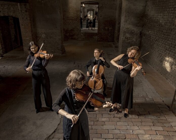 Das Bild zeigt das Chaos String Quartet mit ihren Instrumenten: Die Violinistinnen Susanne Schäffer und Eszter Kruchió, den Cellisten Bas Jongen und die Violistin Sara Marzadori, die sich allesamt in einem aus Backstein-konstruierten Raum befinden