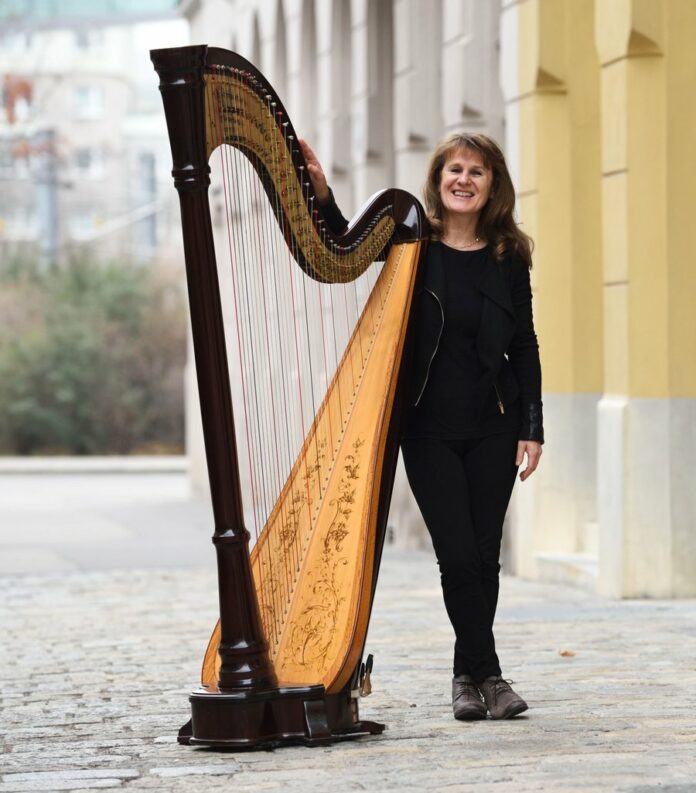 Das Bild zeigt Monika Stadler in Ganzaufnahme, wie sie leicht an ihre Harfe lehnt und freundlich in die Kamera lächelt.