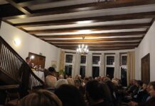 Echtzeitalter – Lesung und Autorengespräch / Botschaft Oslo Das Bild zeigt den Autor Tonio Schachinger im voll besetzten Foyer der österreichischen Botschaft in Oslo. Schachinger sitzt an einem Tisch, der sich am Fuße der Wendeltreppe befindet, und spricht von dort zum darunter sitzenden Publikum