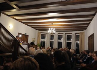 Echtzeitalter – Lesung und Autorengespräch / Botschaft Oslo Das Bild zeigt den Autor Tonio Schachinger im voll besetzten Foyer der österreichischen Botschaft in Oslo. Schachinger sitzt an einem Tisch, der sich am Fuße der Wendeltreppe befindet, und spricht von dort zum darunter sitzenden Publikum