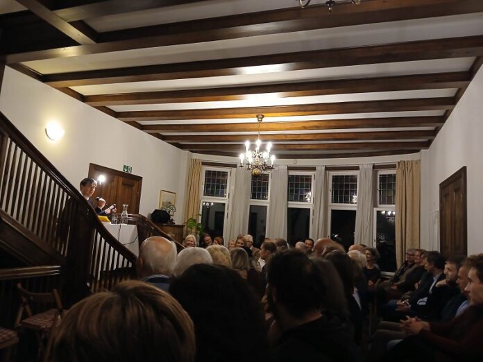 Das Bild zeigt den Autor Tonio Schachinger im voll besetzten Foyer der österreichischen Botschaft in Oslo. Schachinger sitzt an einem Tisch, der sich am Fuße der Wendeltreppe befindet, und spricht von dort zum darunter sitzenden Publikum