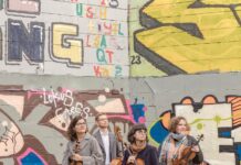 Chaos String Quartet / Kulturforum Ottawa Das Bild zeigt das Chaos String Quartet im Vordergrund, jedes Mitglied mit seinem Instrument in der Hand. Im Hintergrund erstreckt sich eine mit Graffiti bedeckte Wand. Die Mitglieder des Quartetts blicken gemeinsam in dieselbe Richtung – nach rechts.