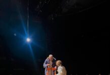 Turrini in Rustawi / Botschaft Tiflis Das Bild zeigt eine Schauspielerin, einen Schauspieler und im Hintergrund die Zuschauerinnen und Zuschauer. Die Schauspielerin steht in der Mitte der Bühne, neben ihr steht der Schauspieler auf der Treppe. Herbstblätter liegen auf der Bühne. Im Vordergrund des Bildes liegt ein Wasserkrug mit einem Herbstblatt darauf auf einem kleinen Tisch.