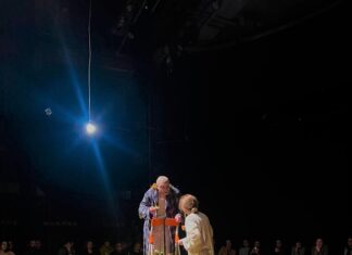Turrini in Rustawi / Botschaft Tiflis Das Bild zeigt eine Schauspielerin, einen Schauspieler und im Hintergrund die Zuschauerinnen und Zuschauer. Die Schauspielerin steht in der Mitte der Bühne, neben ihr steht der Schauspieler auf der Treppe. Herbstblätter liegen auf der Bühne. Im Vordergrund des Bildes liegt ein Wasserkrug mit einem Herbstblatt darauf auf einem kleinen Tisch.
