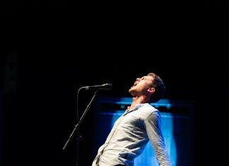 Poetry Slam World Cup Togo/ Botschaft Abuja Das Bild zeigt Emil Kaschka. Er steht auf der Bühne vor einem Mikrofon, mit Hintergrundbeleuchtung. Er trägt ein helles Hemd und eine braune Hose und ist mit dem Oberkörper nach hinten gebeugt und holt mit den Armen aus und ruft dabei etwas ins Mikrofon.