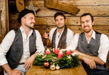 Cobario „Wiener Weihnacht“ / Kulturforum Berlin Das Bild zeigt die drei lächelnden Musiker der Band Cobario in einer gemütlichen, rustikalen Holzhütte. Sie sitzen um einen Holztisch, auf dem ein Adventskranz mit grünen Zweigen, vier brennenden roten Kerzen und weihnachtlichen Dekorationen, wie goldenen und roten Kugeln, liegt. Die Männer tragen weiße Hemden und Westen in gedeckten Farben; einer von ihnen hat einen schwarzen Hut auf. Der Mann in der Mitte hält eine Geige in der Hand. Die Atmosphäre wirkt warm und festlich.