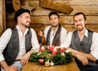 Cobario „Wiener Weihnacht“ / Kulturforum Berlin Das Bild zeigt die drei lächelnden Musiker der Band Cobario in einer gemütlichen, rustikalen Holzhütte. Sie sitzen um einen Holztisch, auf dem ein Adventskranz mit grünen Zweigen, vier brennenden roten Kerzen und weihnachtlichen Dekorationen, wie goldenen und roten Kugeln, liegt. Die Männer tragen weiße Hemden und Westen in gedeckten Farben; einer von ihnen hat einen schwarzen Hut auf. Der Mann in der Mitte hält eine Geige in der Hand. Die Atmosphäre wirkt warm und festlich.