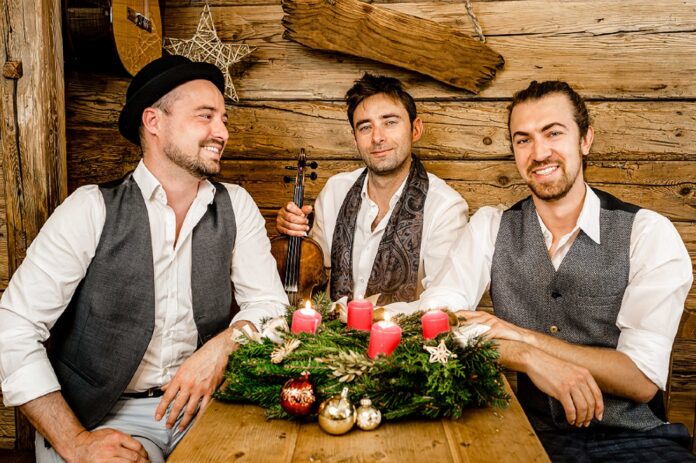 Das Bild zeigt die drei lächelnden Musiker der Band Cobario in einer gemütlichen, rustikalen Holzhütte. Sie sitzen um einen Holztisch, auf dem ein Adventskranz mit grünen Zweigen, vier brennenden roten Kerzen und weihnachtlichen Dekorationen, wie goldenen und roten Kugeln, liegt. Die Männer tragen weiße Hemden und Westen in gedeckten Farben; einer von ihnen hat einen schwarzen Hut auf. Der Mann in der Mitte hält eine Geige in der Hand. Die Atmosphäre wirkt warm und festlich.