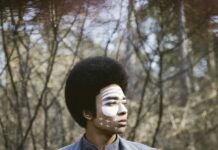 Open Air-Konzert beim „Weihnachtsmarkt in der Altstadt“ / Kulturforum Bratislava Portrait von DJ Kimyan Law: im Hintergrund sind Bäume unscharf zu sehen. Er trägt ein rauchblaues Hemd und eine rauchblaue Jacke. Er hat quer über das Gesicht eine weisse Bemalung und blickt nach rechts aus dem Bild