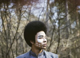 Open Air-Konzert beim „Weihnachtsmarkt in der Altstadt“ / Kulturforum Bratislava Portrait von DJ Kimyan Law: im Hintergrund sind Bäume unscharf zu sehen. Er trägt ein rauchblaues Hemd und eine rauchblaue Jacke. Er hat quer über das Gesicht eine weisse Bemalung und blickt nach rechts aus dem Bild