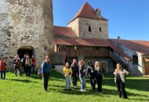 Siebenbürgen Connect / Kulturforum Bukarest Das Bild zeigt die Schülerinnen undSchüler aus Bad Ischl auf einer Reise in Siebenbürgen, Rumänien. Sie stehen vor mittelalterlichen Gebäuden auf einer Wiese, mit blauen Himmel und Sonnenschein.